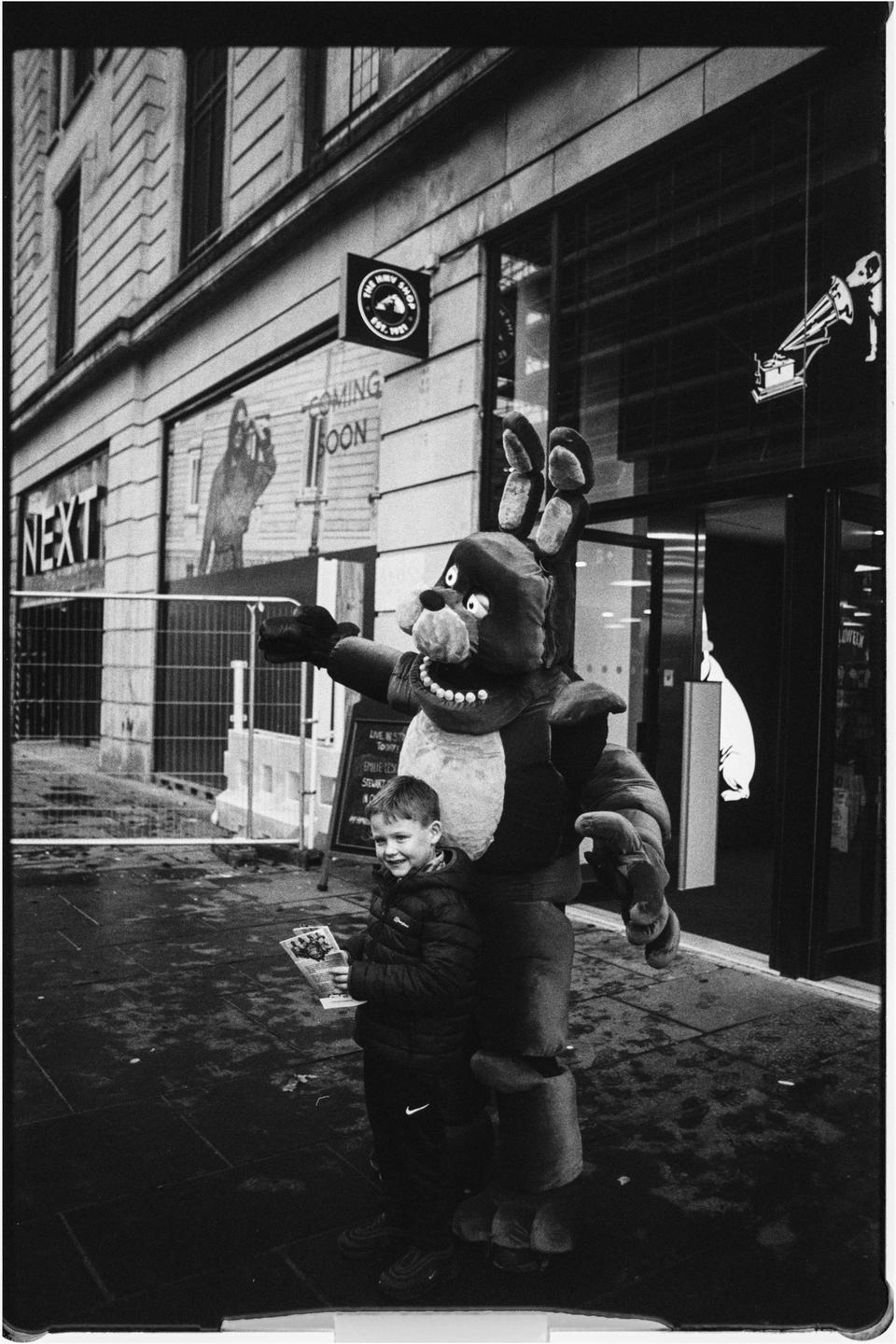 A boy stands next to a person in a large animal costume on a wet street.