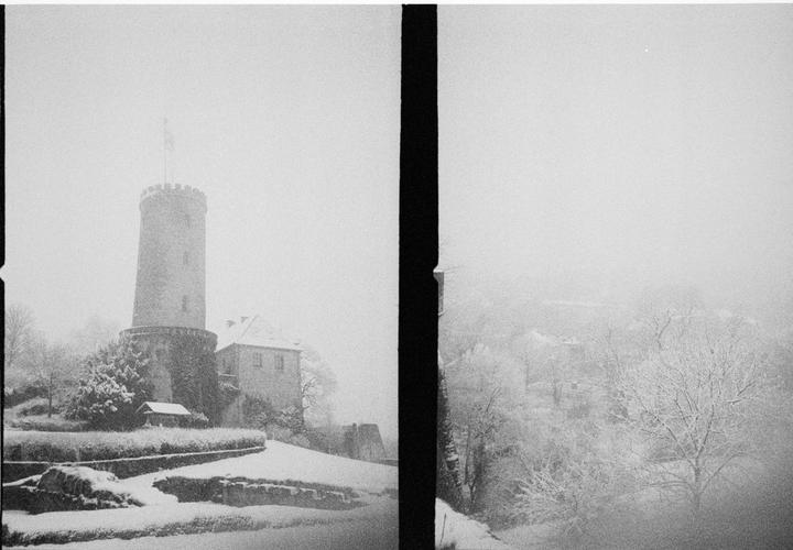 A tall, round tower with a flag on top stands next to a building, surrounded by snow-covered trees and ground in a hazy, winter landscape.