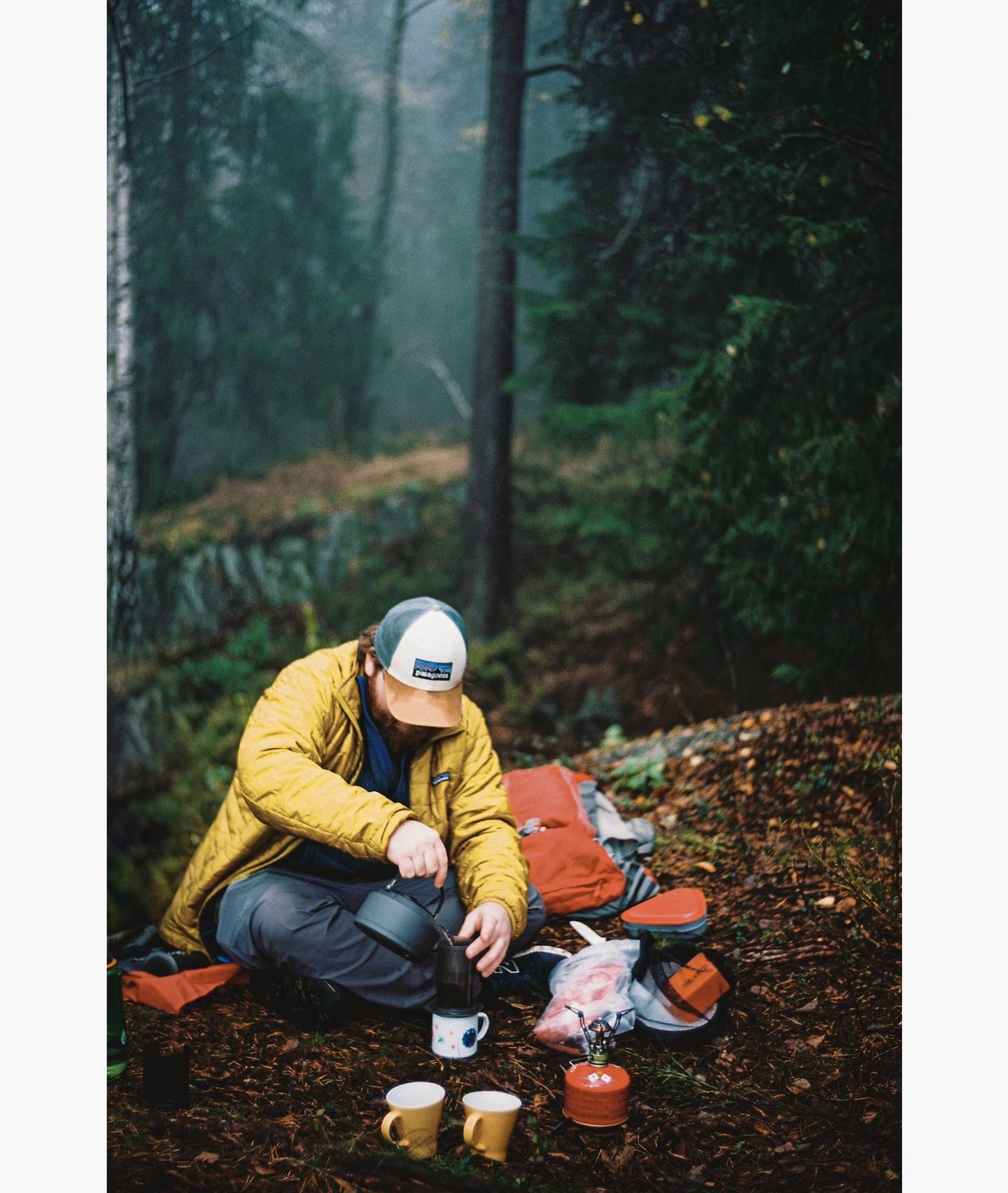 A man wearing a yellow jacket and a cap is preparing coffee in a forest.