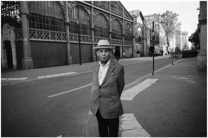 A man in a hat and jacket stands on a street in front of a large building with arched windows.