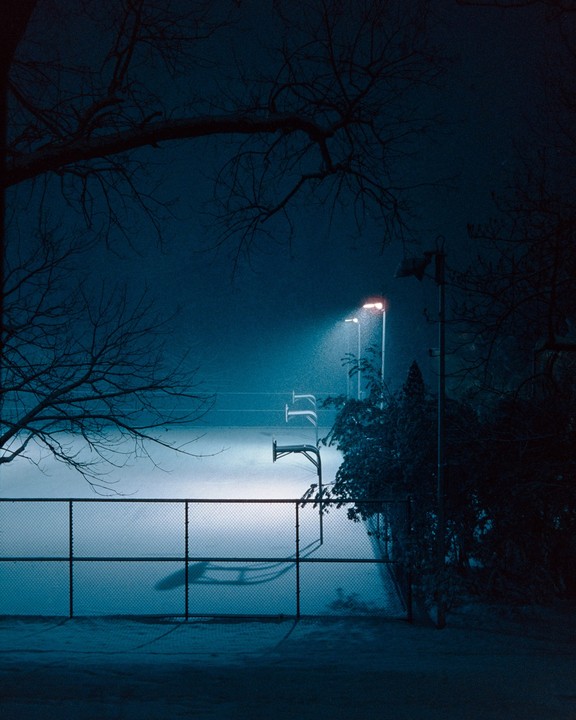 A basketball court is illuminated by a street light at night, with snow covering the ground and bare tree branches framing the top of the image.