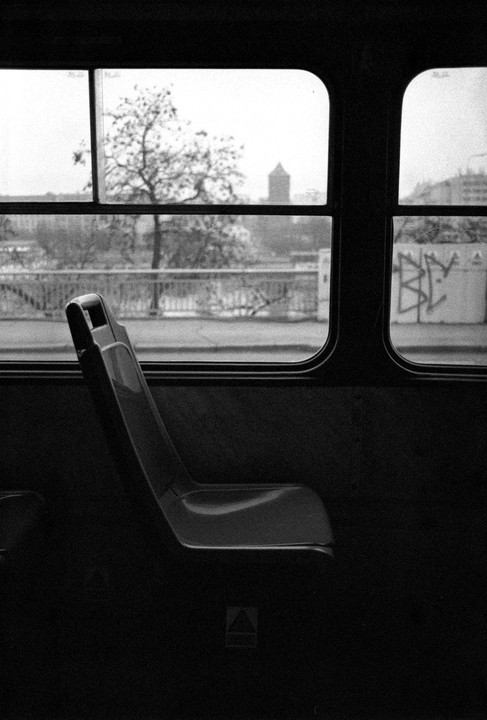A dark, empty seat is visible inside a vehicle, with a view of a bridge, trees, and a distant building through the window.