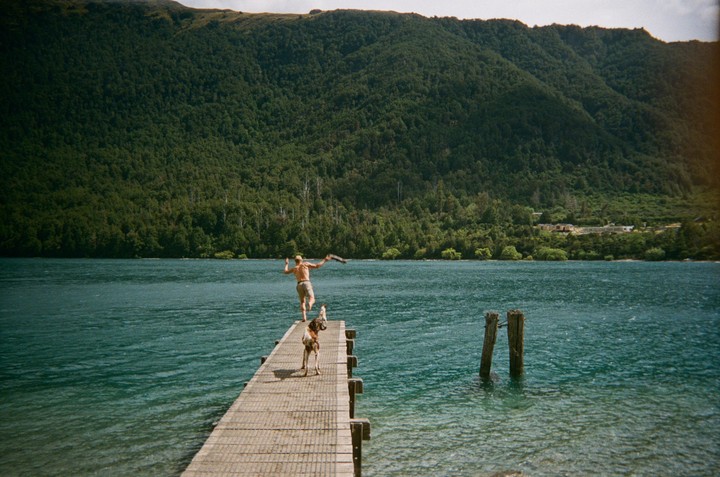 A man throws a stick for a dog from a dock on a lake with a forested mountain in the background.