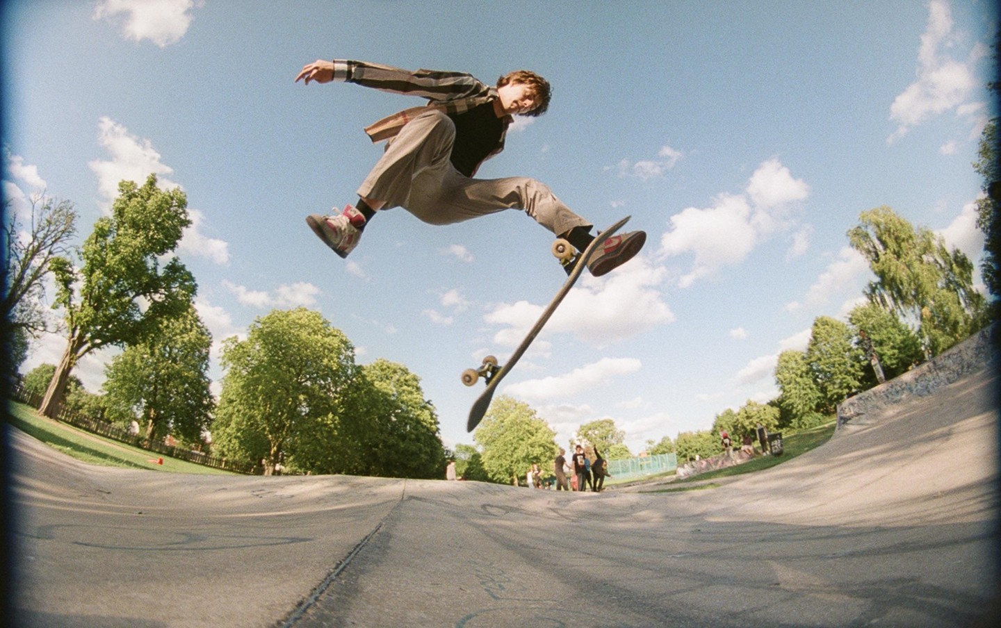 A young man performs a skateboarding trick in a skatepark under a blue sky with scattered clouds.
