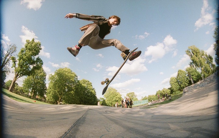 A young man performs a skateboarding trick in a skatepark under a blue sky with scattered clouds.