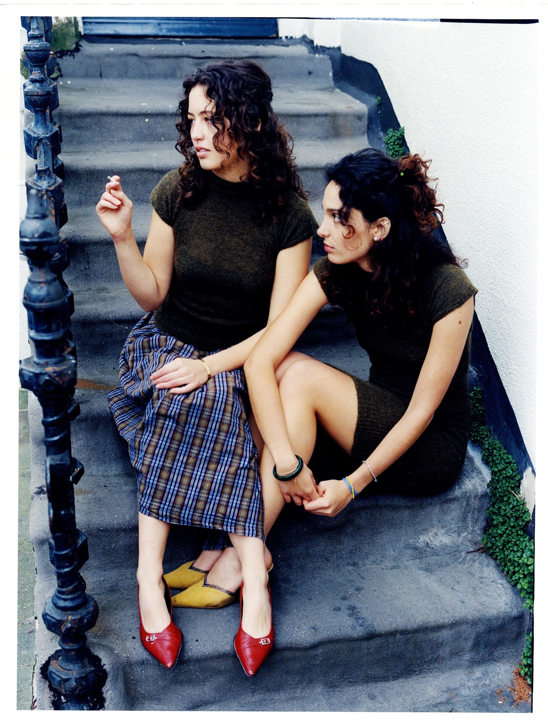Two women with curly hair sit on concrete steps, one holding a cigarette and the other looking to the side.