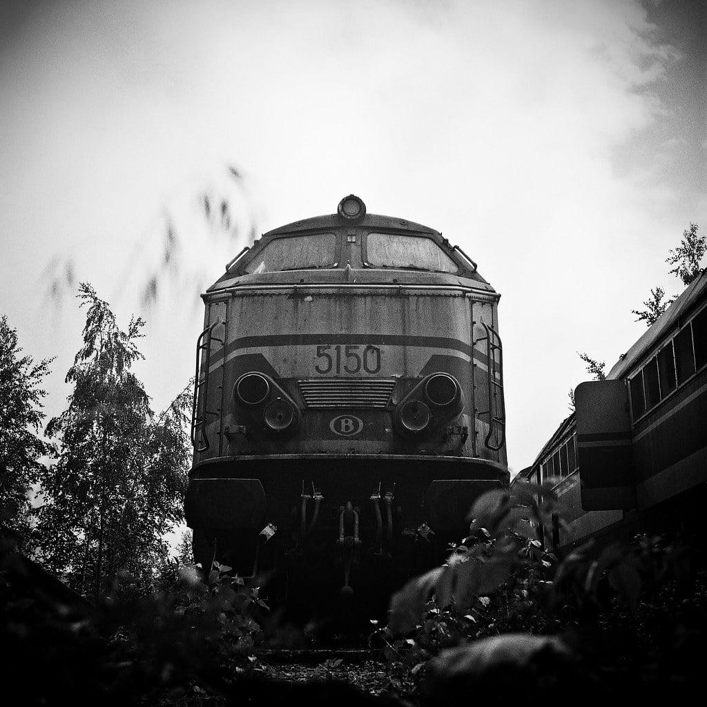 A black and white photograph shows the front of an old train engine with the number 5150 on it, surrounded by overgrown vegetation.