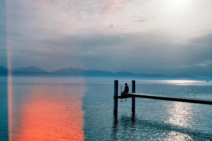 A person sits on a dock over a lake with mountains in the distance under a cloudy sky.