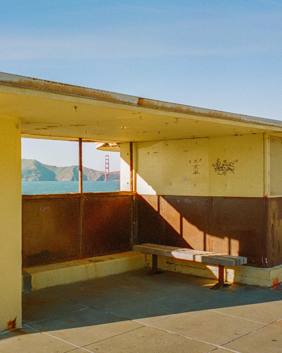 A bench sits in a partially enclosed structure with a view of the Golden Gate Bridge and mountains across the water.