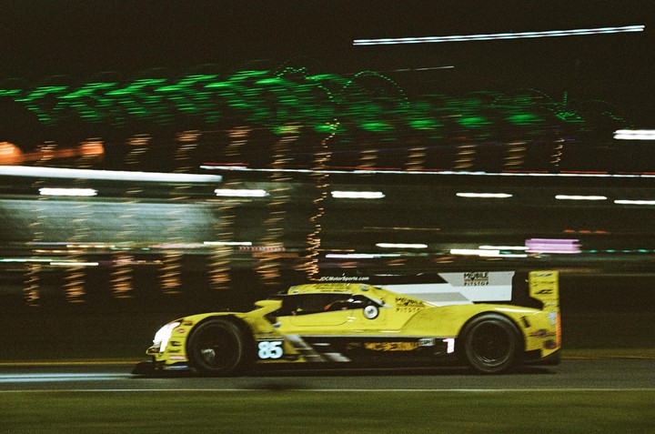 A yellow race car with the number 85 on its side speeds past a blurred background of green and white lights at night.