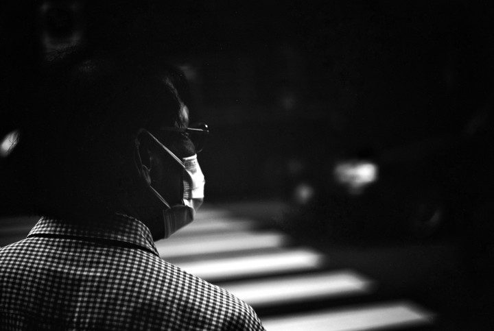 A man wearing a face mask and glasses stands with his back to the viewer, looking towards a crosswalk at night.