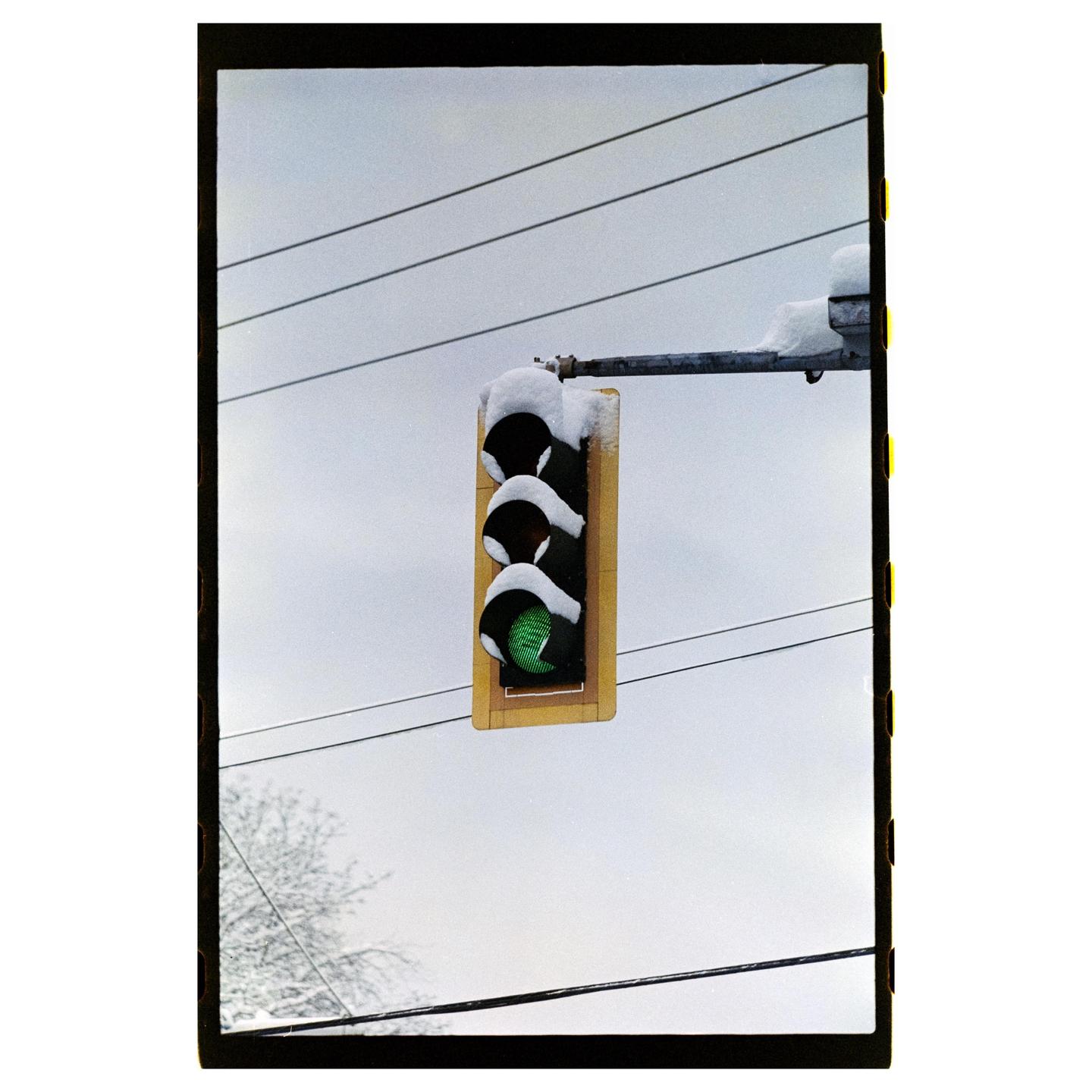A traffic light covered in snow hangs against a cloudy sky with power lines and a snow-dusted tree visible.