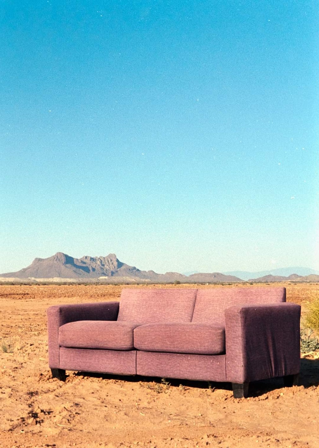 A purple sofa sits in a barren, dry landscape with mountains in the distance under a clear blue sky.
