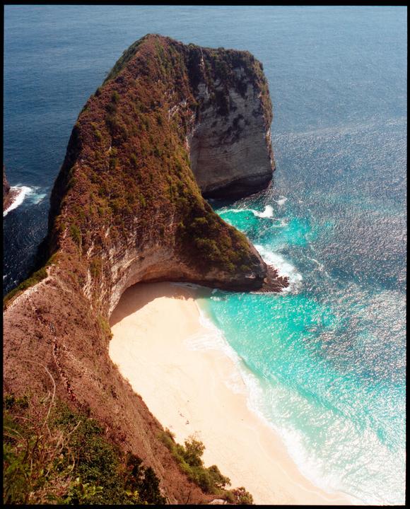 A high-angle view shows a white sand beach and turquoise water at the base of a large, green-covered cliff.