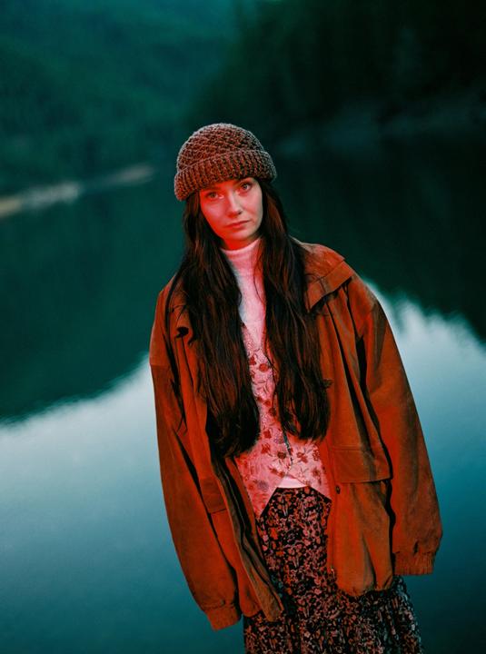 A woman wearing a knit hat and a brown jacket stands in front of a body of water.