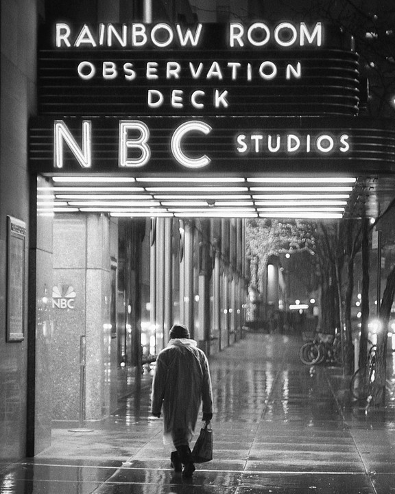A person in a raincoat walks away from the illuminated NBC Studios sign on a wet city street at night.