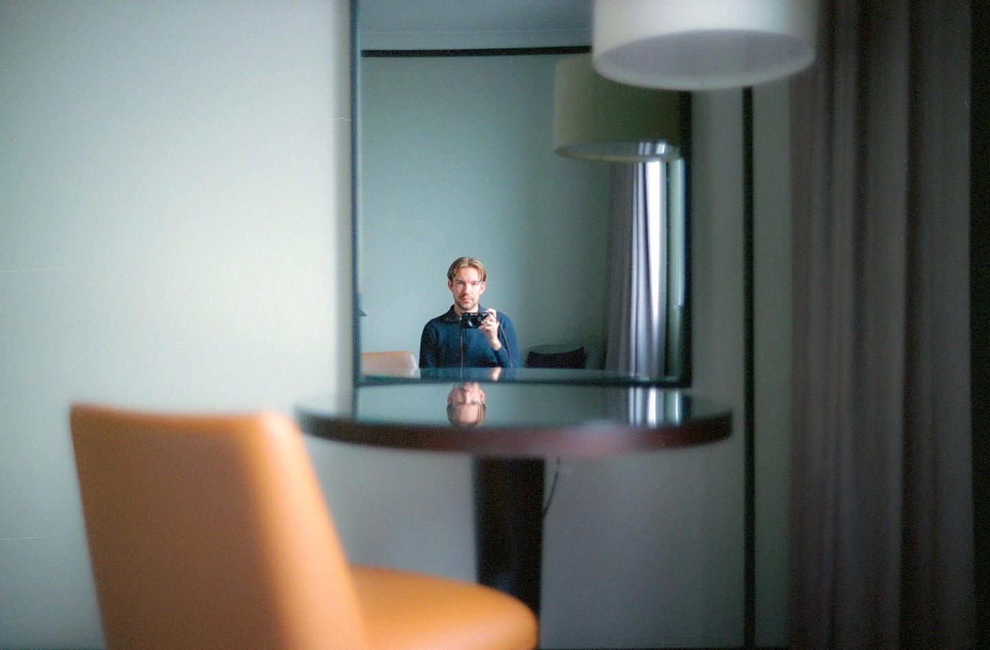 A man with a camera is reflected in a mirror above a round table with a chair in the foreground.