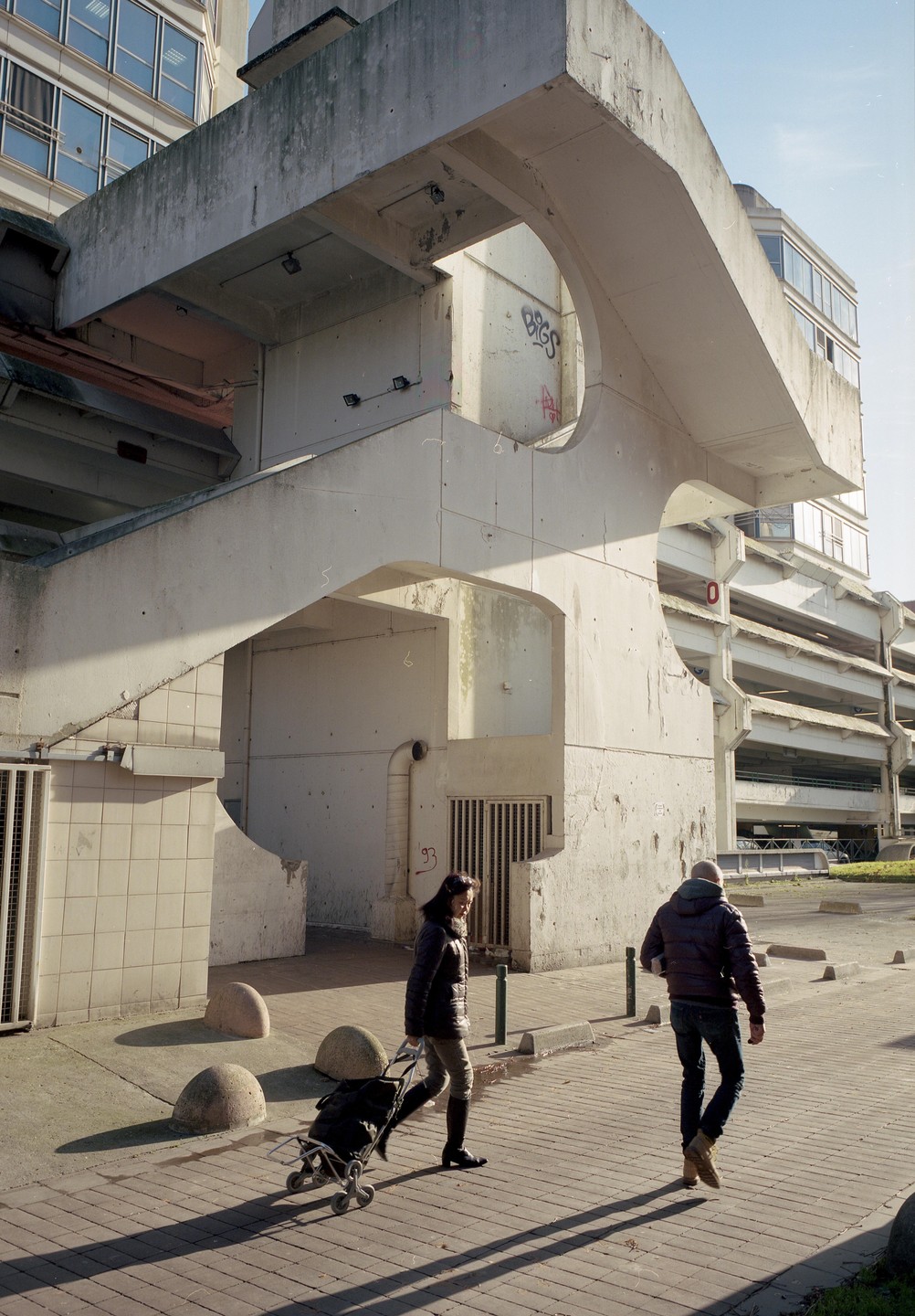 A woman pulls a shopping cart past a man walking on a paved street next to a concrete building.
