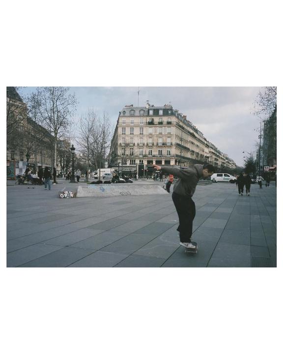 A man on a skateboard balances in a city square with buildings and trees in the background.