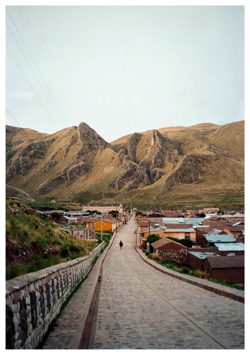 A person walks down a cobblestone street towards a village nestled in a valley with mountains in the background.