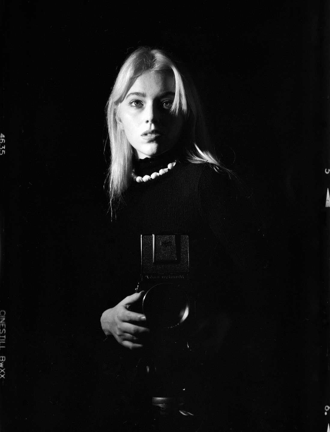 A woman with blonde hair and a pearl necklace holds a camera in a self-portrait.