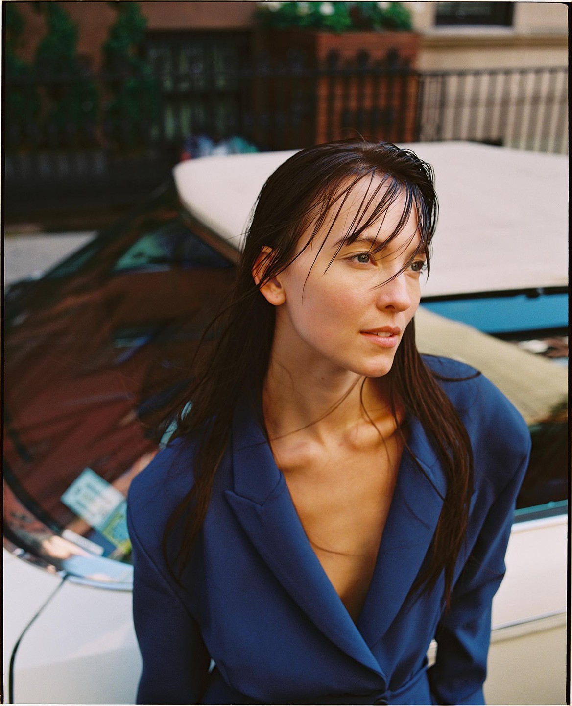 A woman with wet hair and a blue jacket looks to the side while leaning on a car.
