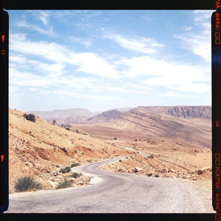 A winding road cuts through a desert landscape with mountains in the background under a cloudy sky.