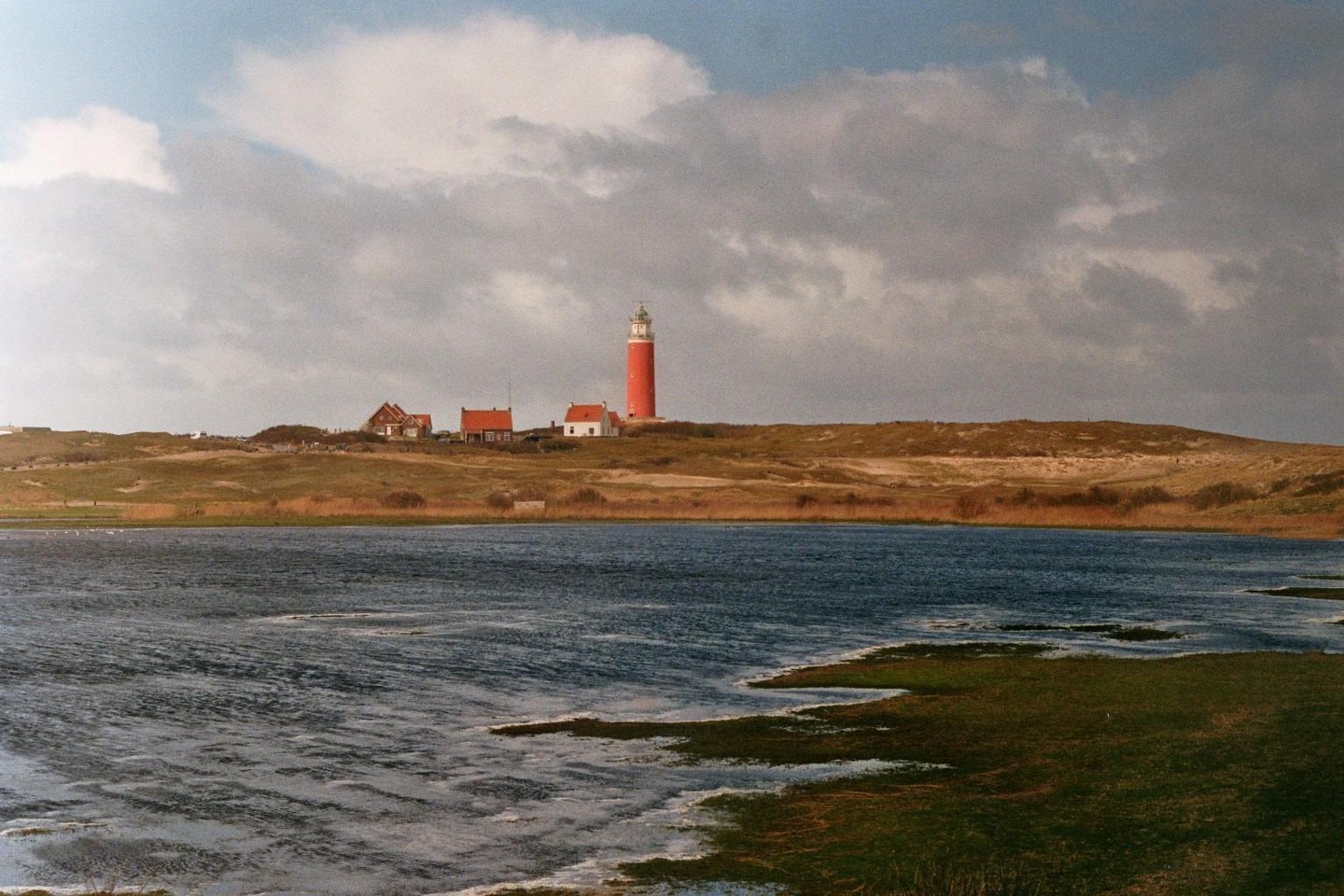 A red lighthouse stands on a grassy hill overlooking a body of water with small houses nearby under a cloudy sky.