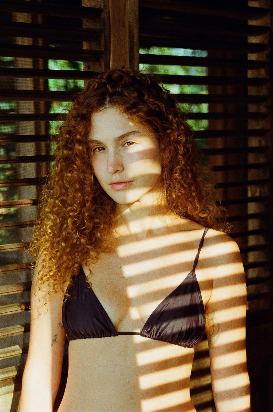 A woman with curly red hair stands in a bikini top with shadows from blinds across her body.