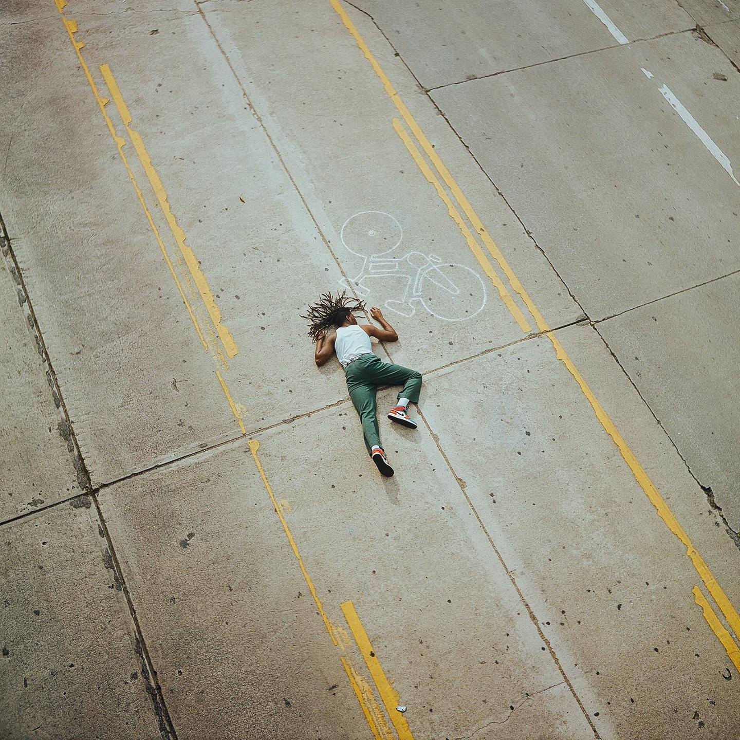 A person with dreadlocks lies on a concrete road next to a chalk drawing of a bicycle.
