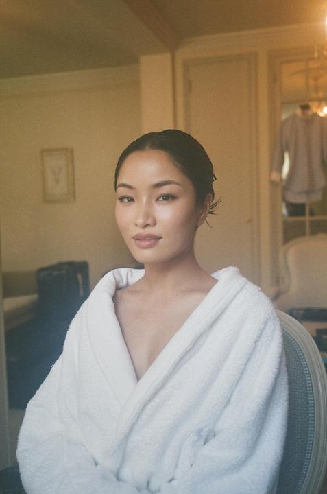 A woman in a white robe sits in a room with a chair and a framed picture in the background.