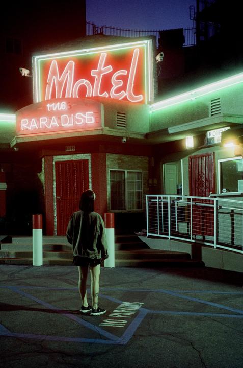 A woman stands in a parking lot looking at a motel with neon signs at night.
