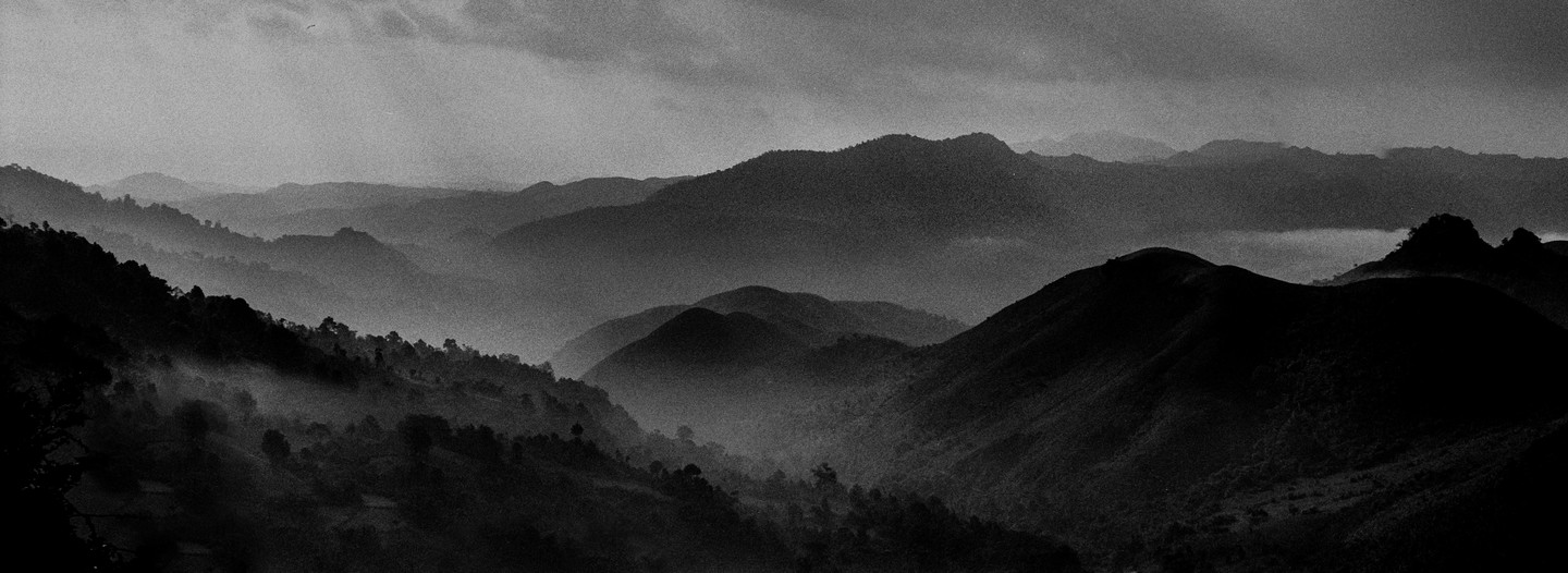 A black and white landscape shows layers of mountains and hills covered in mist under a cloudy sky.