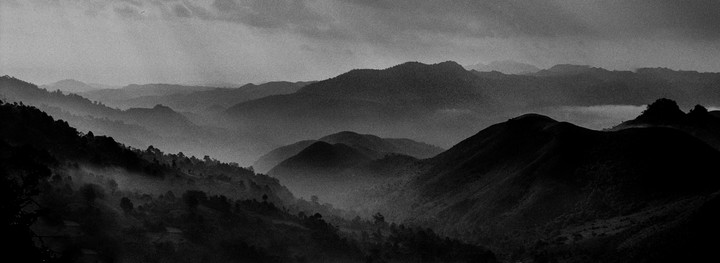 A black and white landscape shows layers of mountains and hills covered in mist under a cloudy sky.