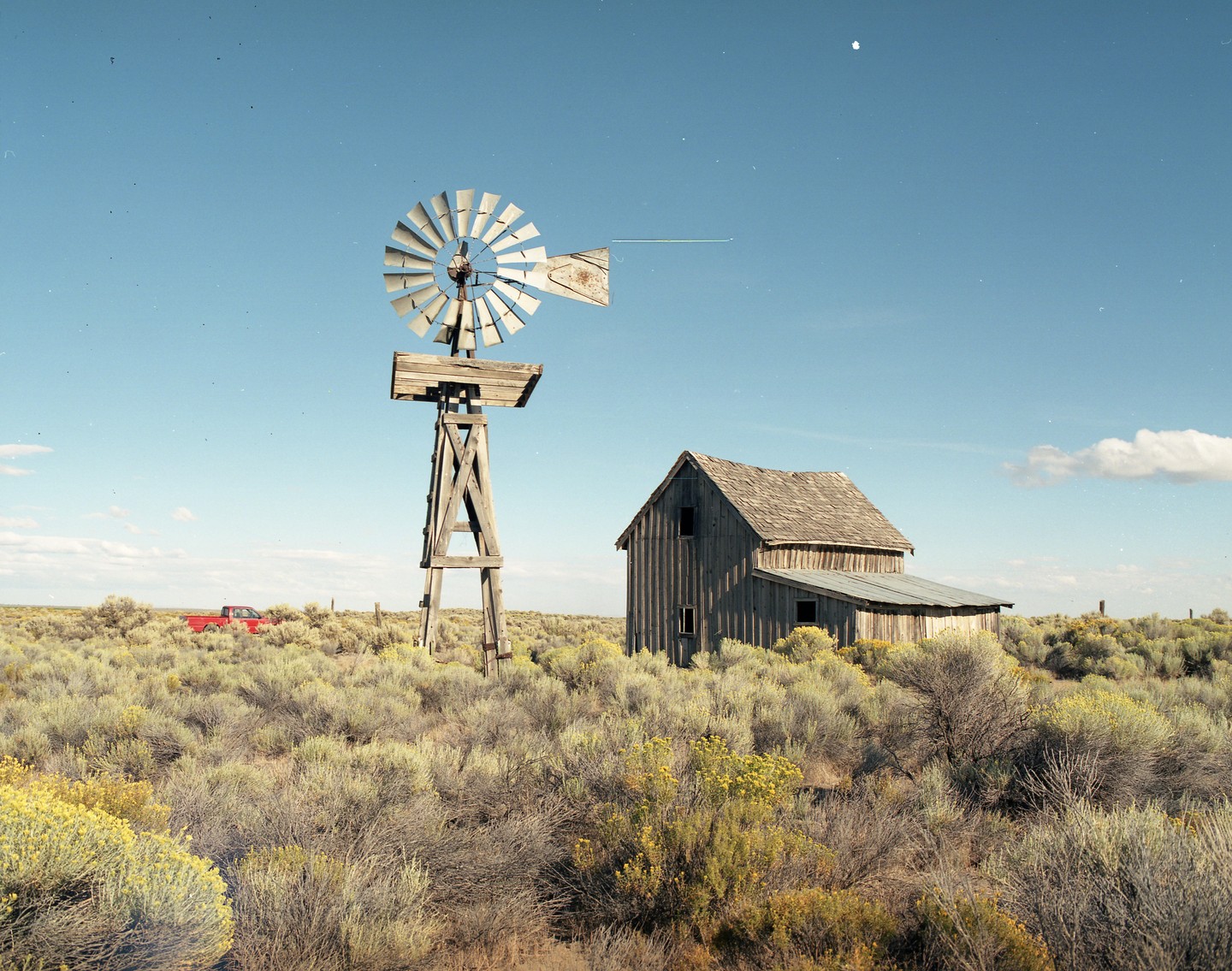 A wooden windmill and an old wooden house stand in a field of sagebrush under a clear sky.