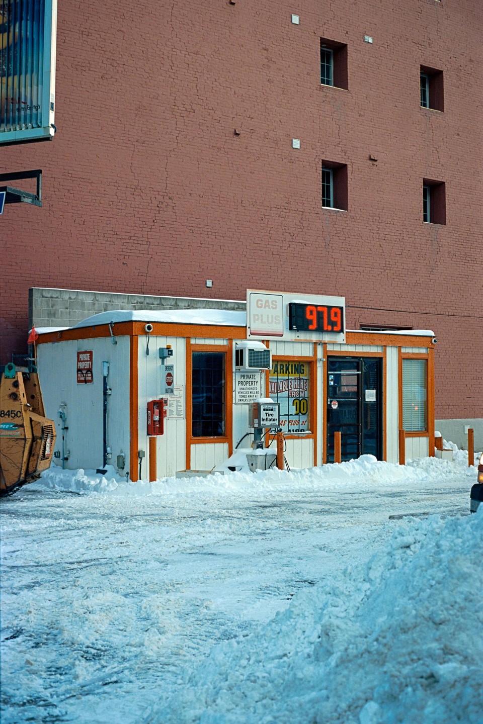 A small gas station building with an orange trim is surrounded by snow, with a large brick building in the background.