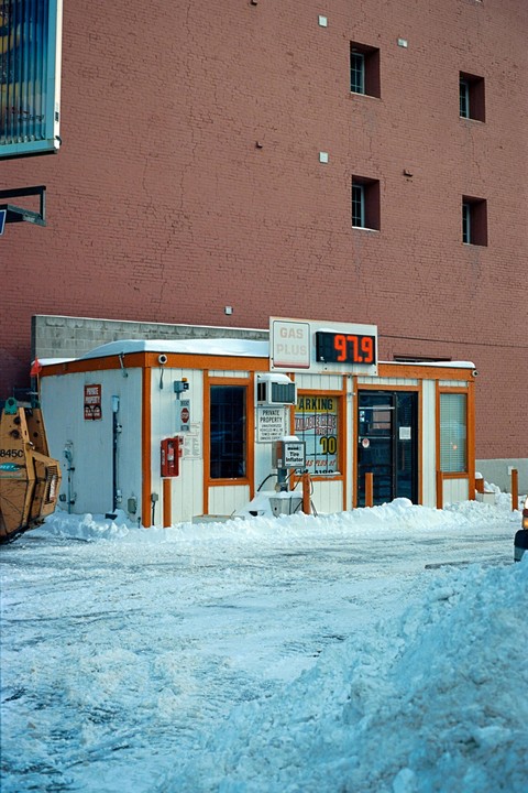 A small gas station building with an orange trim is surrounded by snow, with a large brick building in the background.
