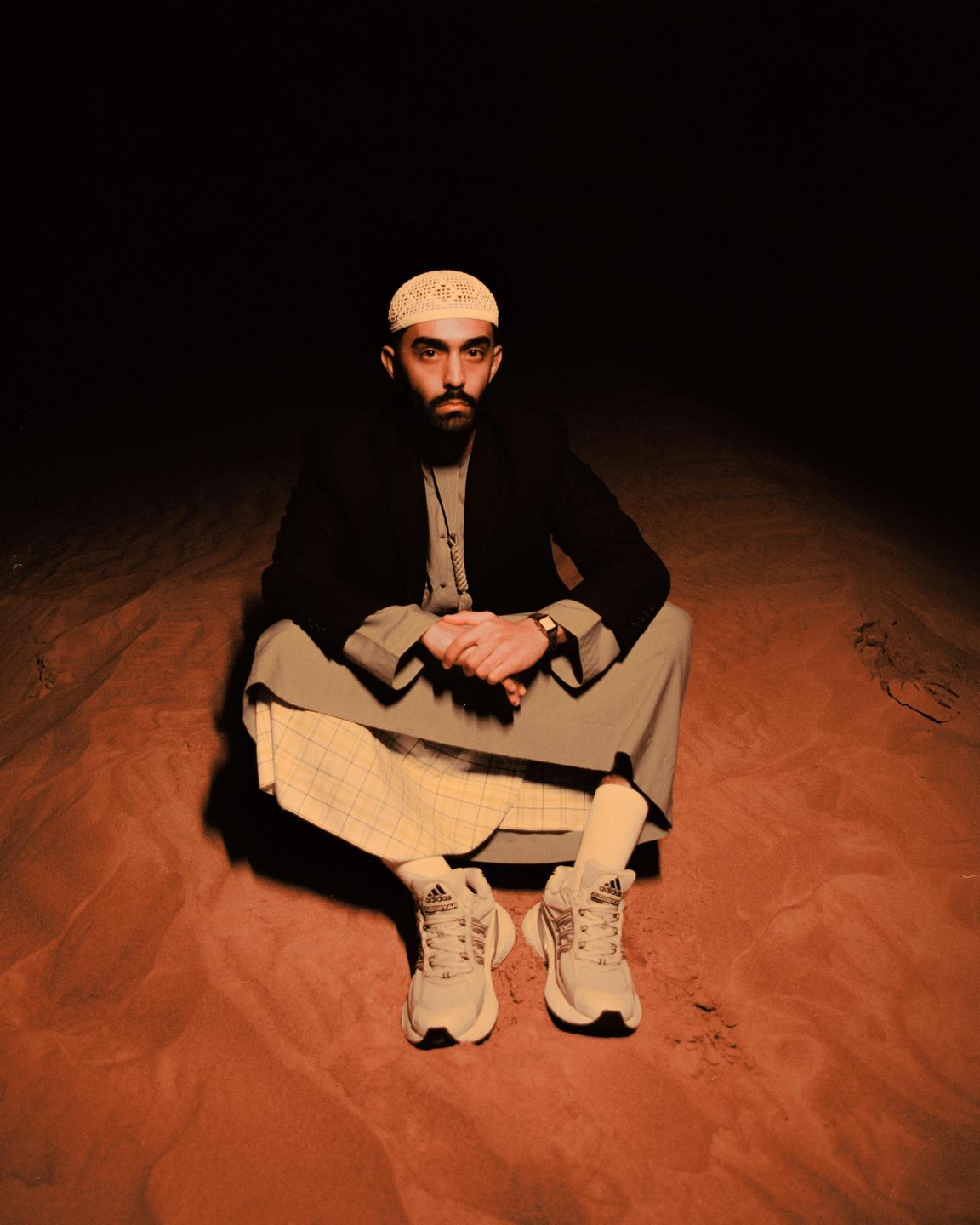 A man wearing a kufi, blazer, and thobe sits on sand in a dark setting.