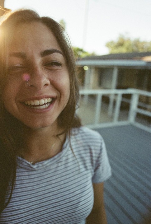 A woman with a nose ring is laughing with her eyes closed, with a deck and house in the background.