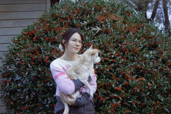 A woman holds a small dog in front of a large bush with red berries.