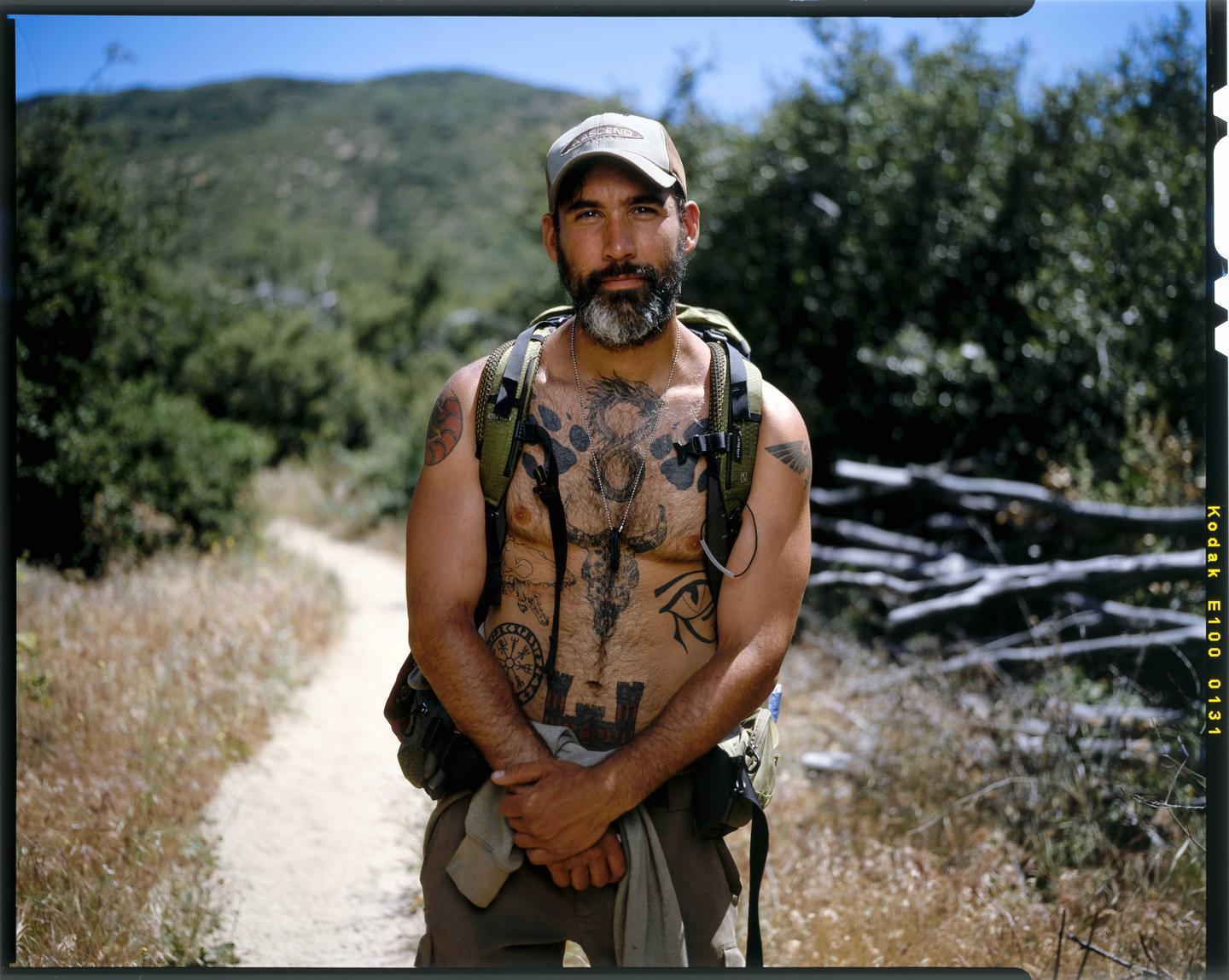 A man with a beard and tattoos stands on a dirt path with a backpack, surrounded by dry grass and green foliage.
