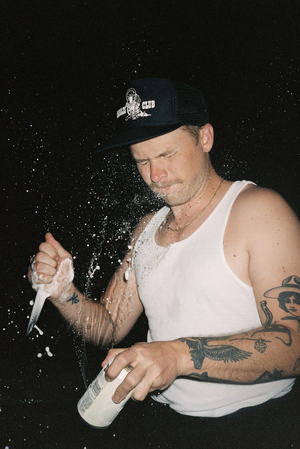 A man in a tank top and hat sprays liquid from a can onto his face and arm, with a knife in his other hand.