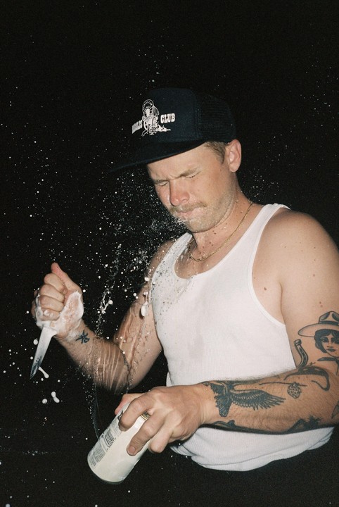 A man in a tank top and hat sprays liquid from a can onto his face and arm, with a knife in his other hand.