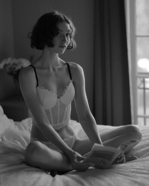 A woman in lingerie sits on a bed and holds a book.