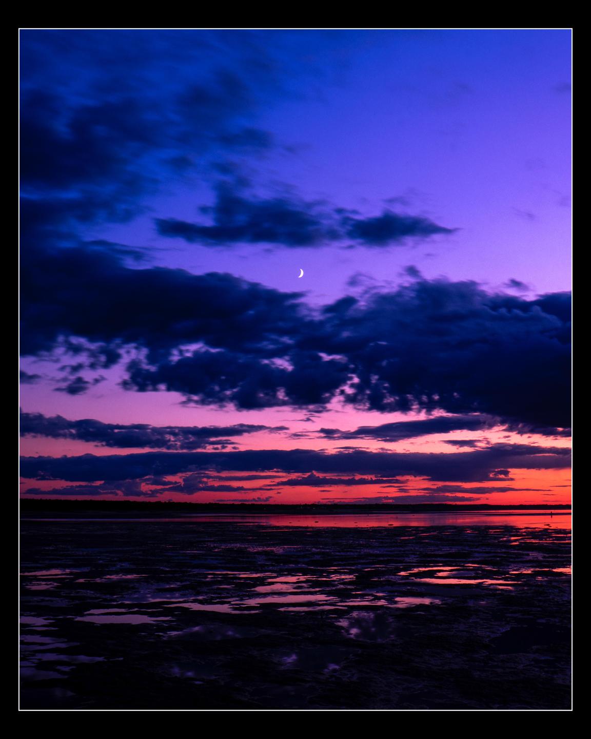 A crescent moon is visible in the purple and red sky over a body of water at sunset.