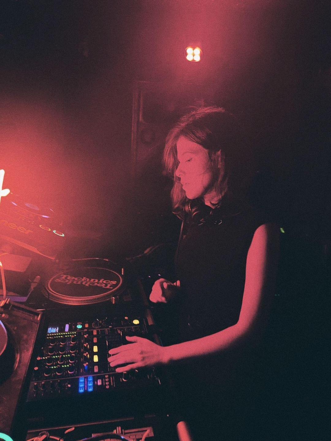 A woman with long hair stands in a dark club, illuminated by red light, with her hands on a DJ mixer.