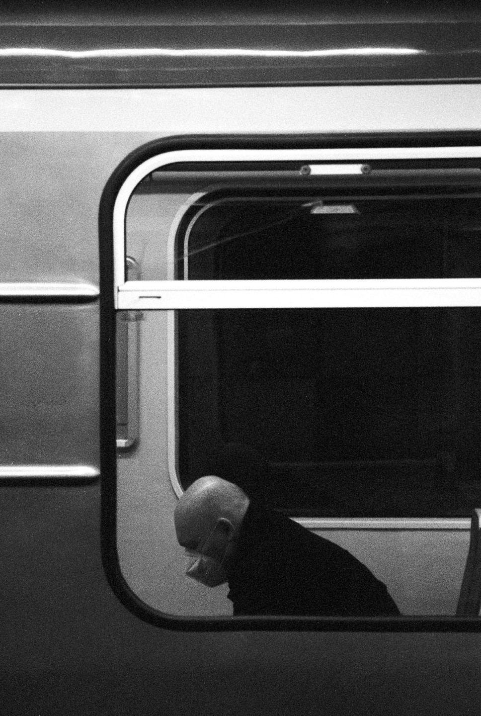 A bald man wearing a face mask is visible through the window of a train.