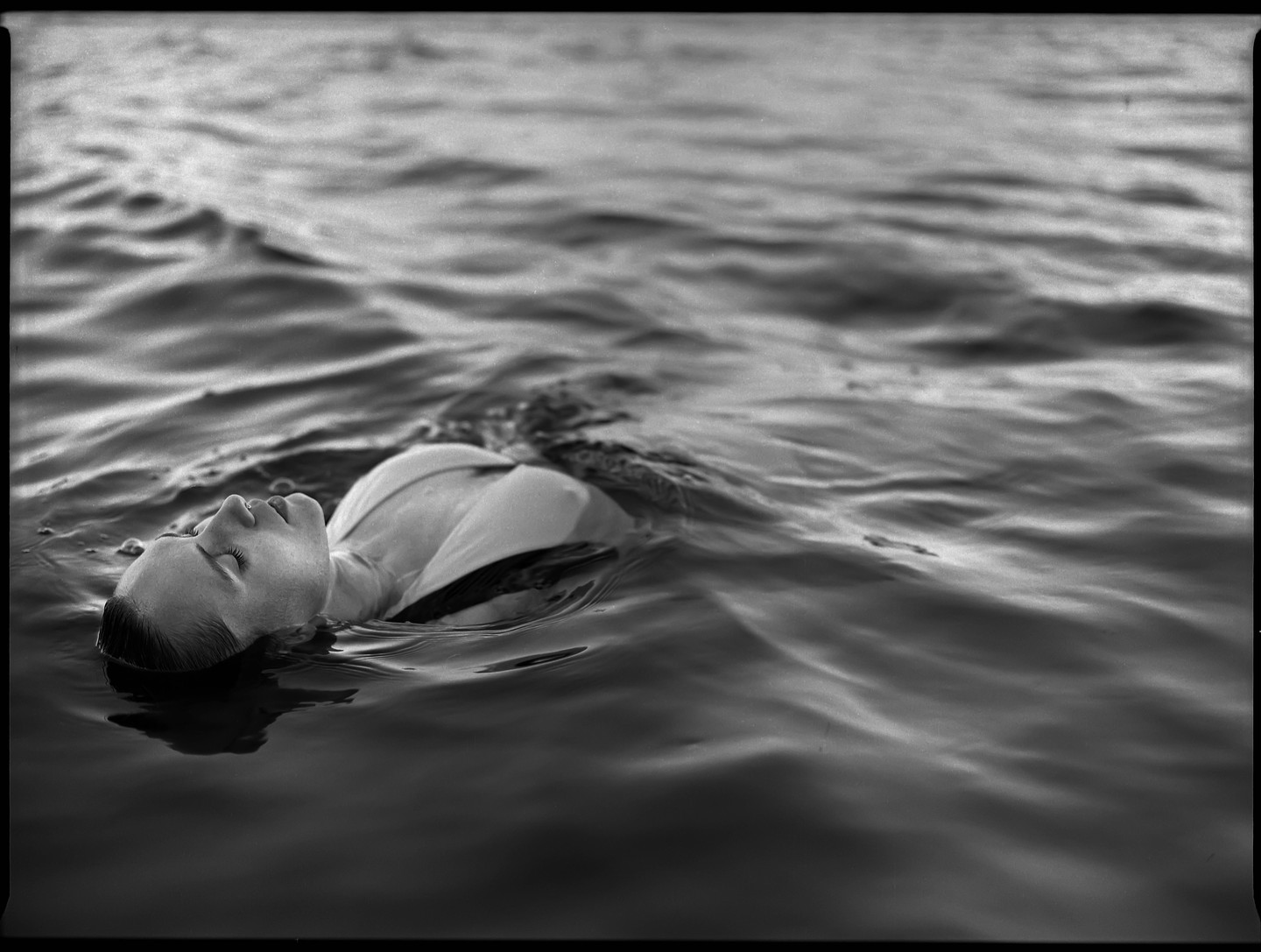 A woman floats on her back in rippling water with her eyes closed.