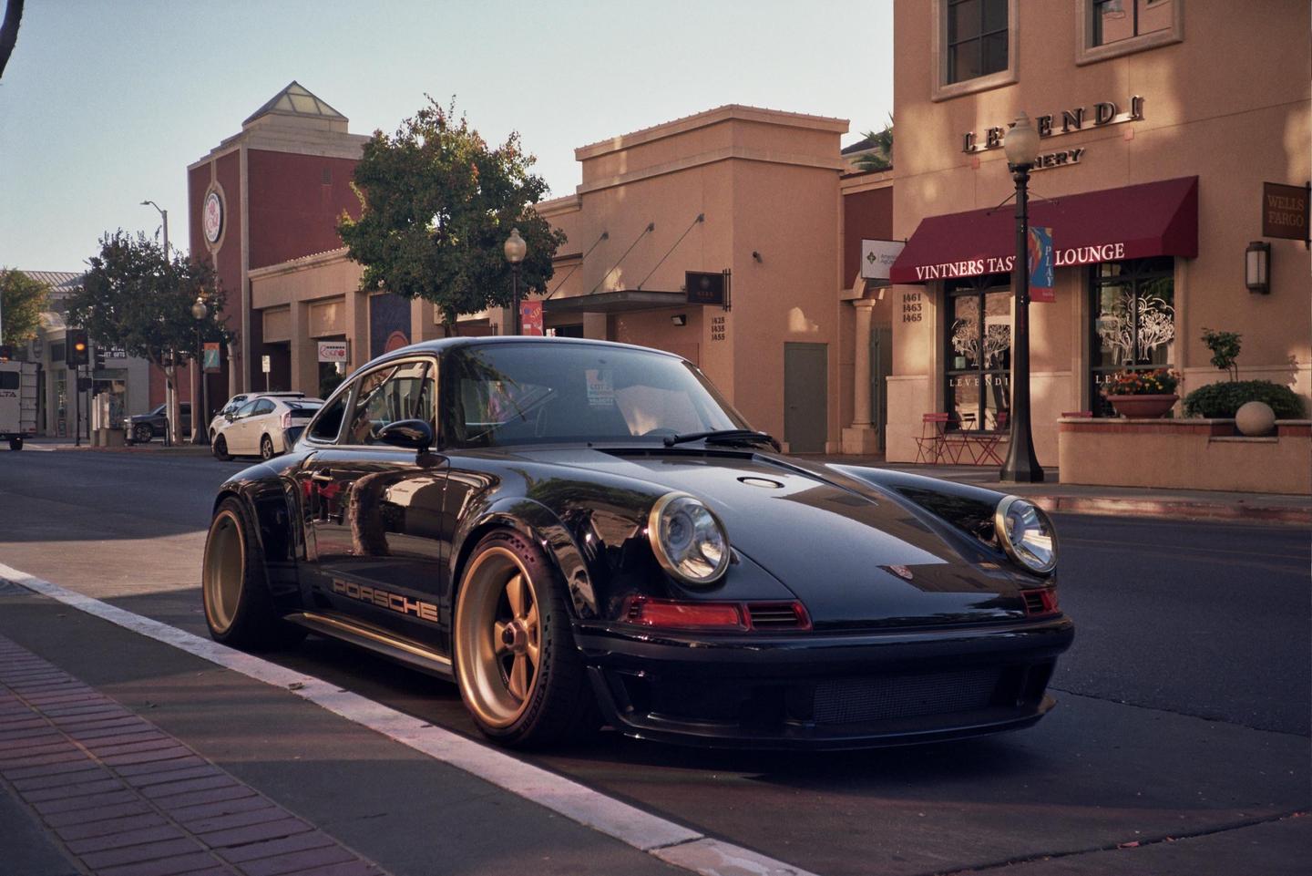 A black Porsche with gold wheels is parked on a street in front of buildings with awnings.