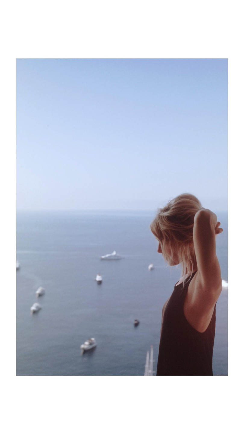 A woman with her hair up looks out over the sea with boats in the water.